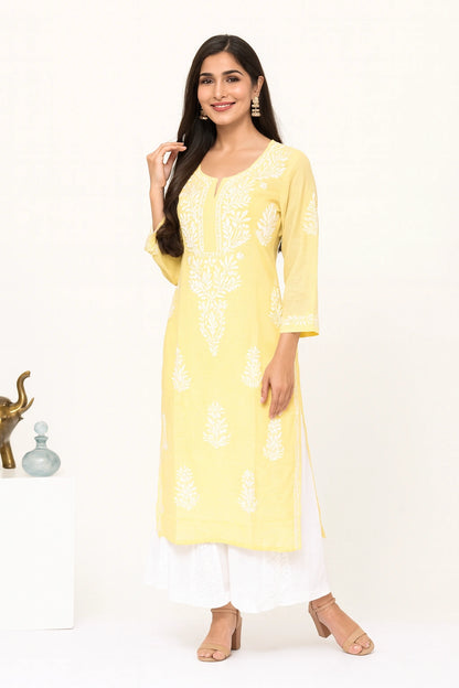 Lucknowi chikankari A woman modeling a light yellow kurta with white pants and silver sandals in a bright, modern studio setting with decorative white pedestals.