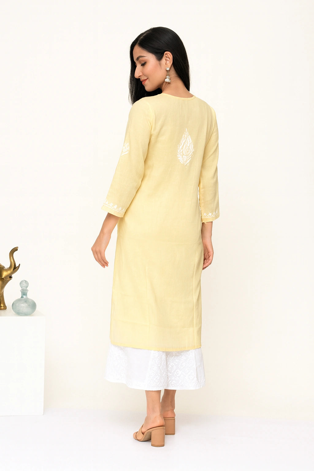 A woman wearing a light yellow embroidered kurta with white pants and beige block-heel sandals, shown from the back in a minimalist indoor setting.