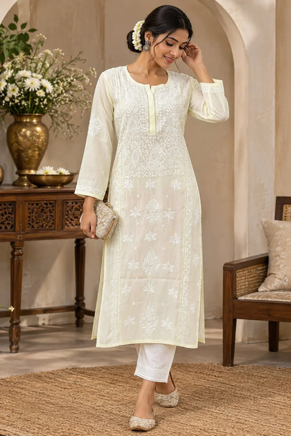 A woman models a light yellow kurta with intricate white embroidery, paired with white cropped pants, in an outdoor courtyard setting decorated with flowers and wooden furniture.