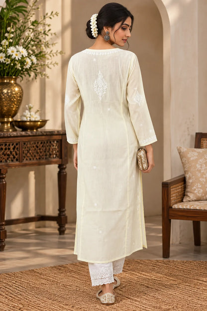 A woman wearing a yellow-colored embroidered kurta with white pants, standing in front of a carved wooden door with floral decorations.