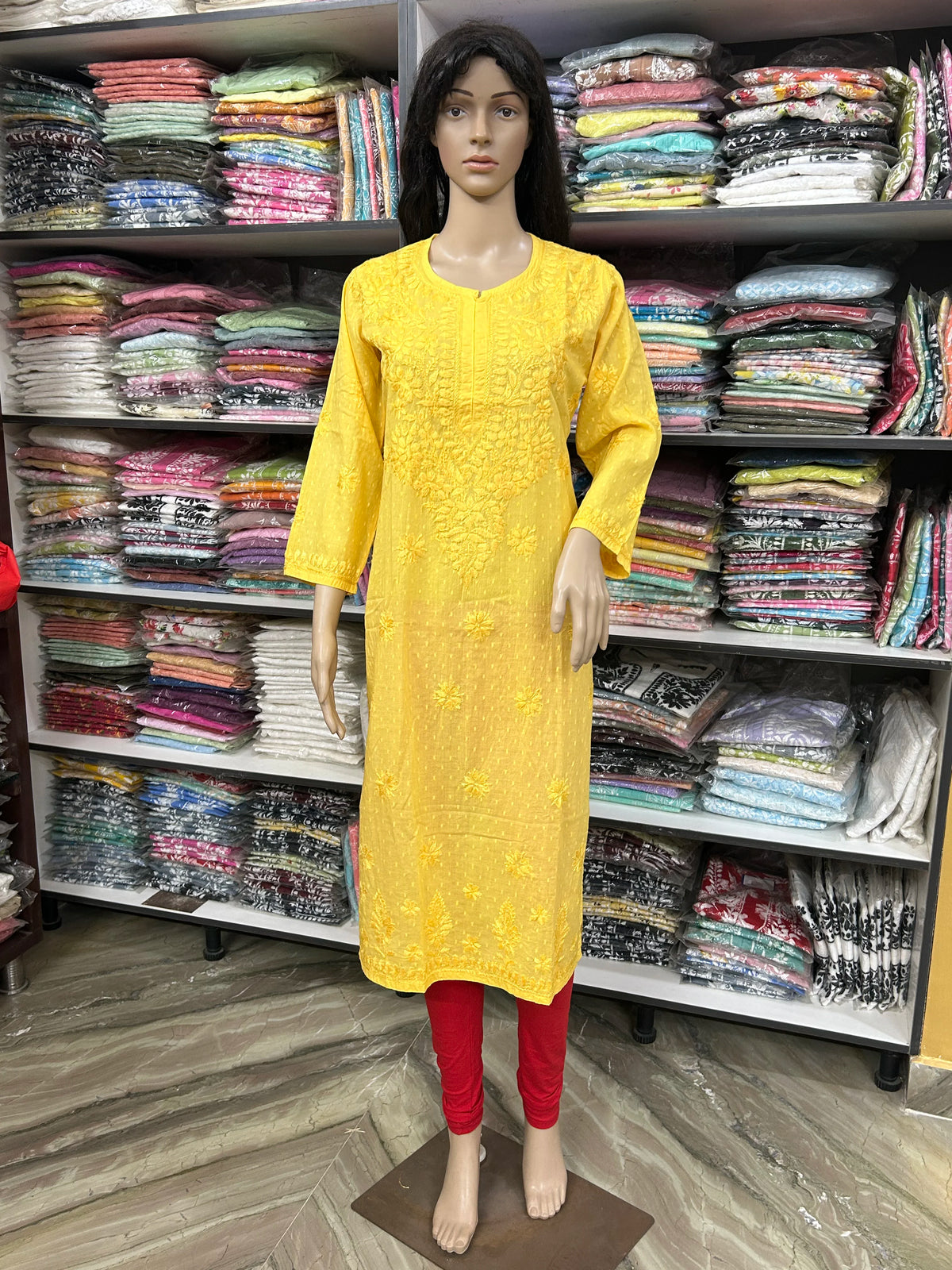 A yellow embroidered kurta displayed on a female mannequin, paired with red leggings, set against a background of shelves filled with folded colorful garments.