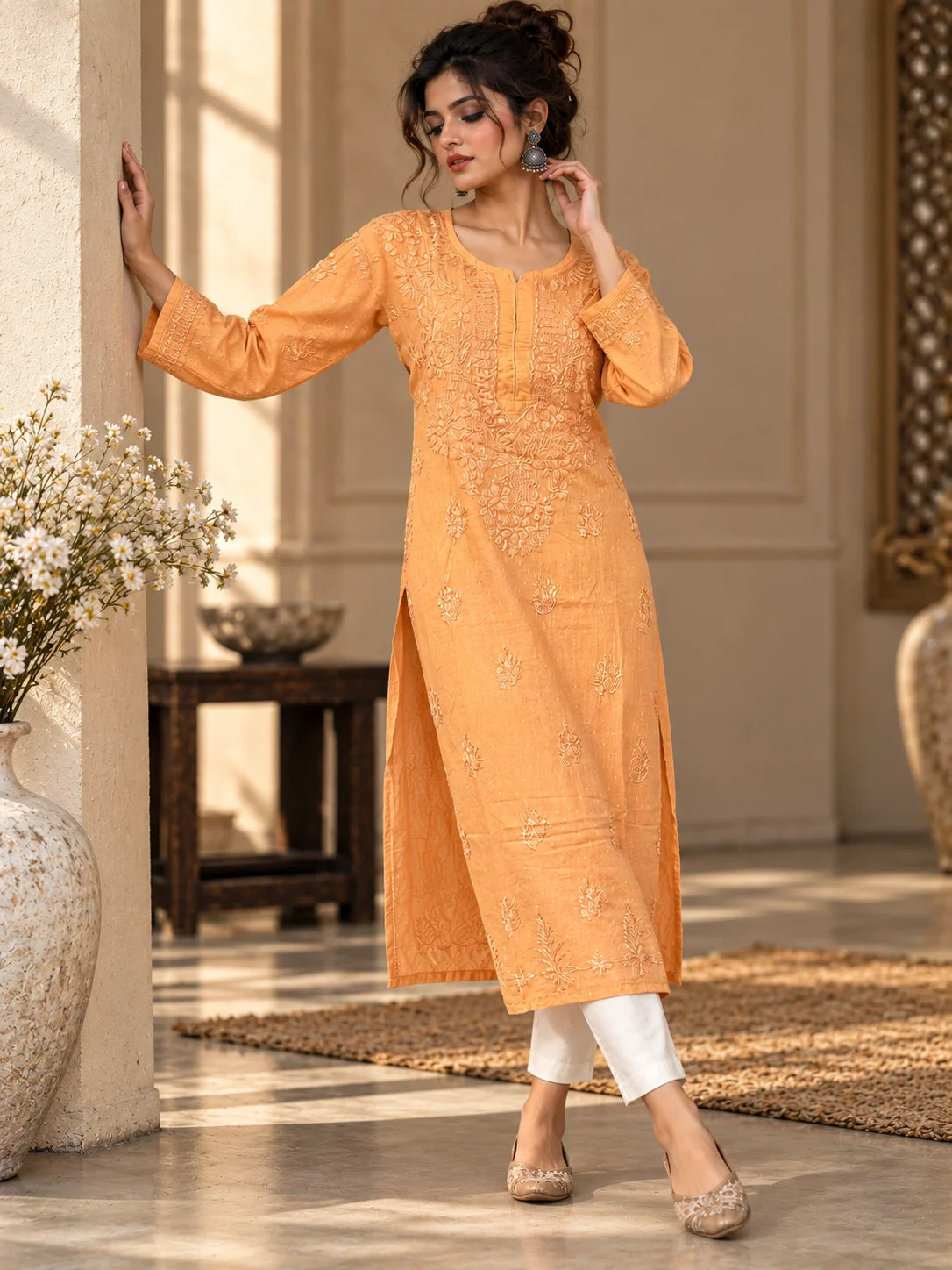A woman models a peach-orange embroidered kurta with white pants and beige shoes in a warm, sunlit indoor setting with classical architecture.