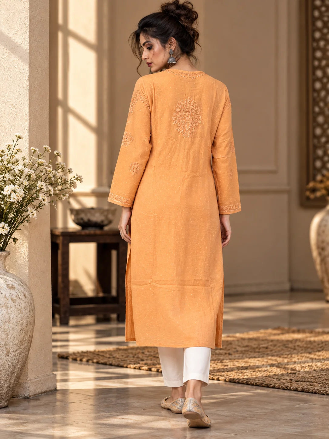 A woman wearing a peach-orange cotton kurta with embroidered details on the back and sleeves, paired with white pants and beige flat shoes, walking in a sunlit indoor setting.