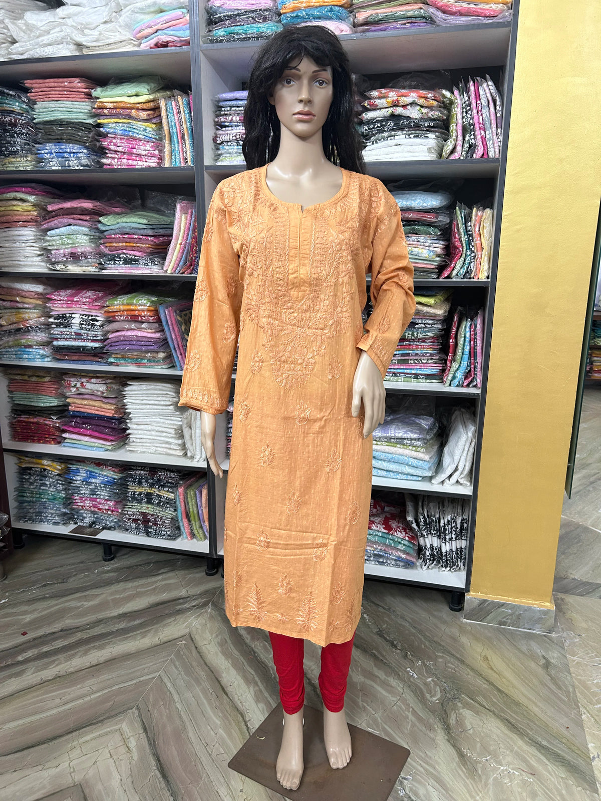 An orange embroidered long kurta displayed on a female mannequin, paired with red pants, set against a background of shelves filled with folded colorful fabrics.