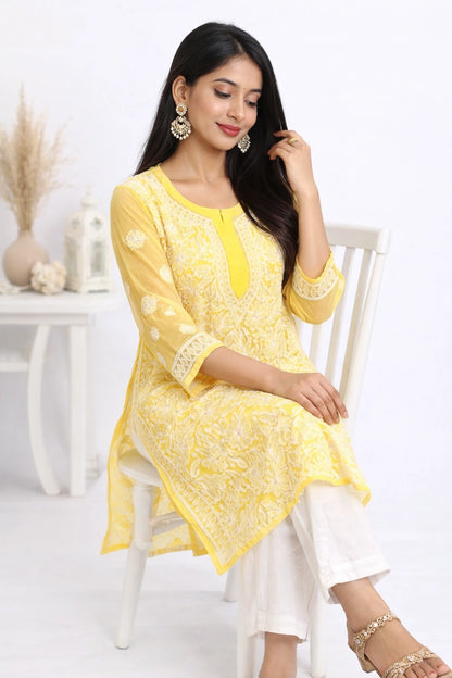 A woman wearing a bright yellow embroidered kurta with white pants, sitting on a white wooden chair in a bright, clean room setting.