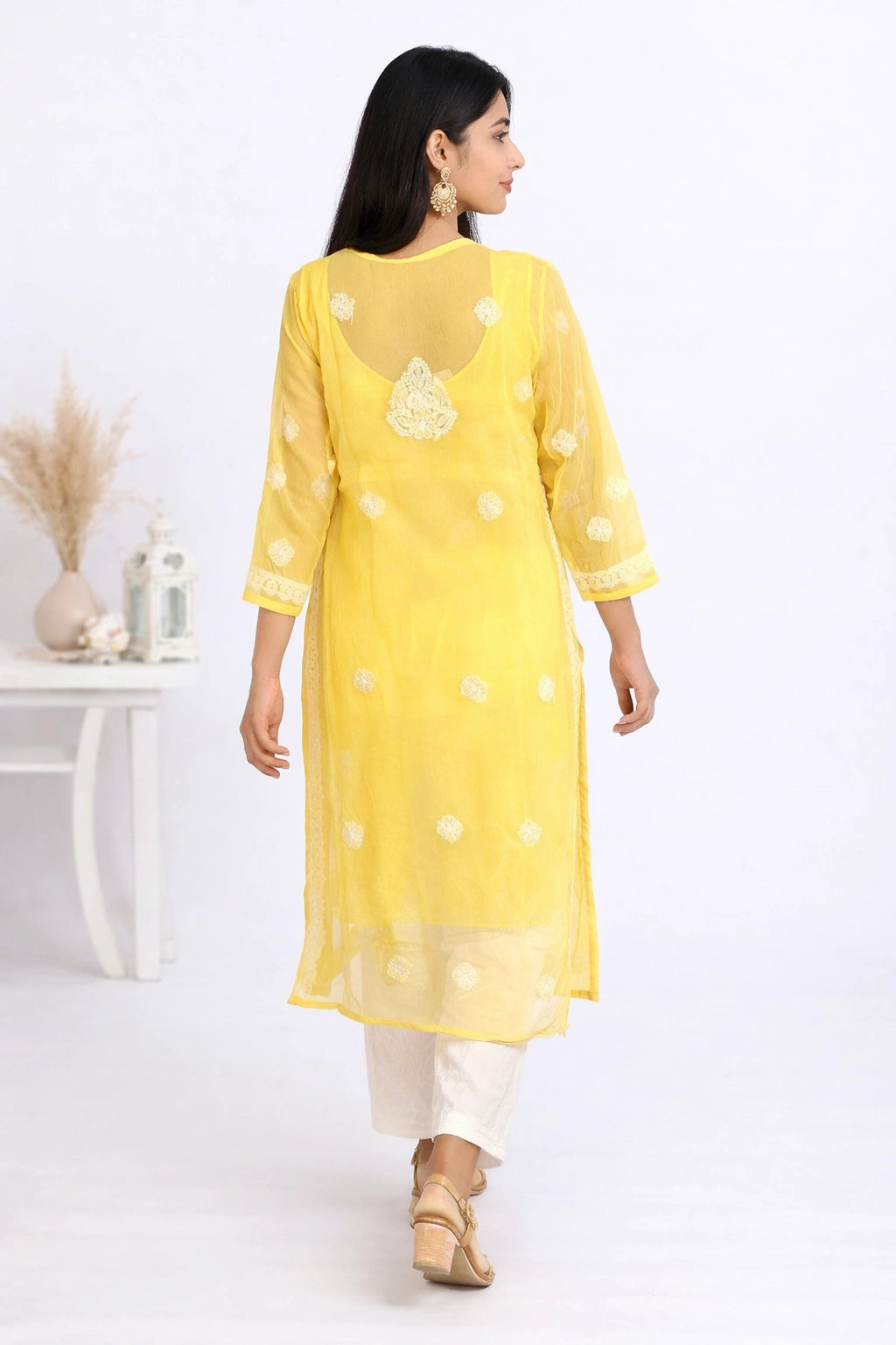 A woman wearing a bright yellow embroidered kurta with white pants and beige sandals, shown from the back in a clean white studio setting.