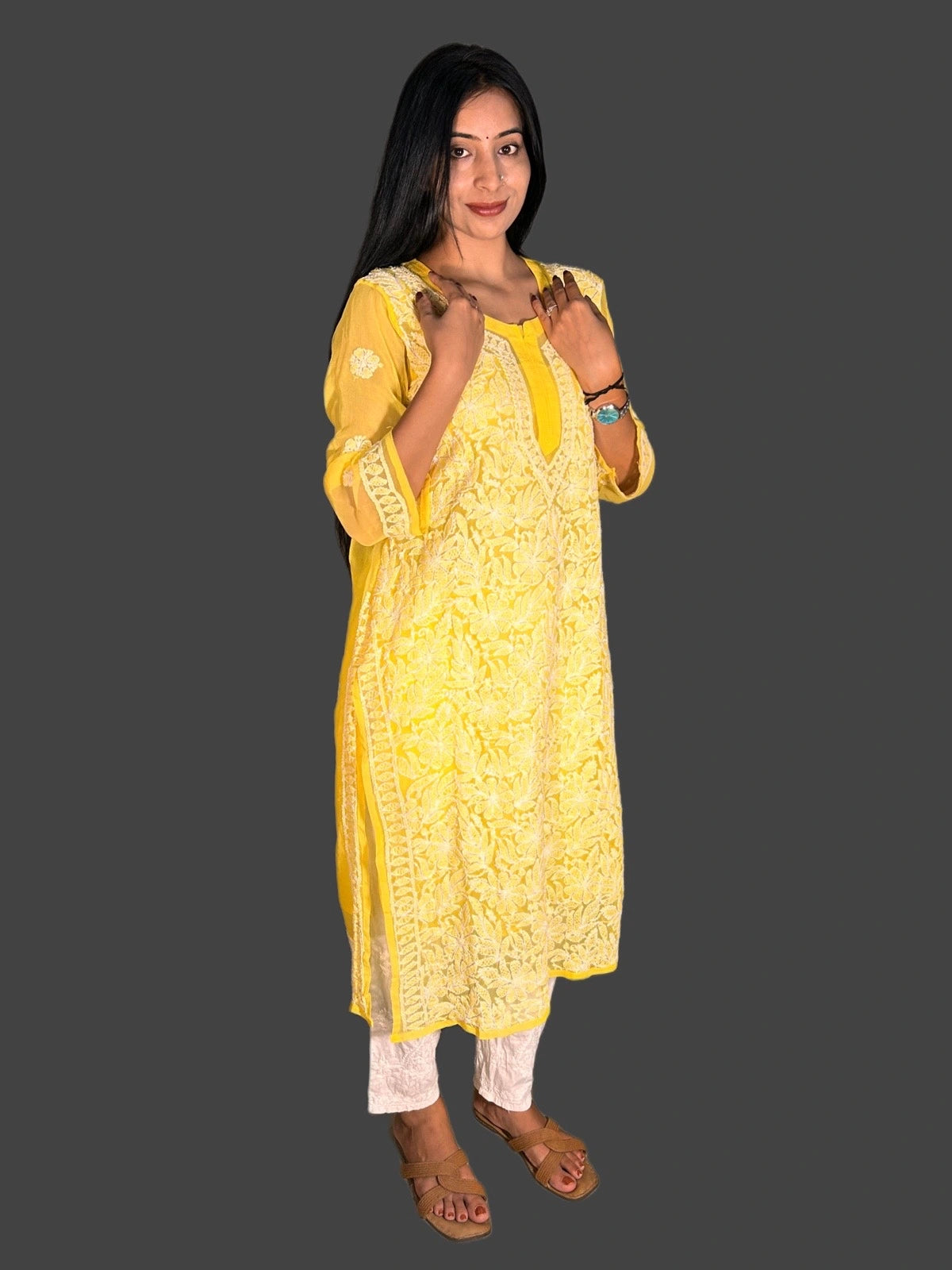 A woman modeling a yellow embroidered kurta with white pants and brown sandals, set against a solid dark gray background.