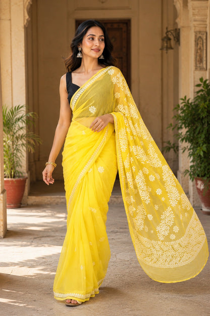 Lucknowi chikankari A woman wearing a bright yellow saree with a black sleeveless blouse, standing in a sunlit courtyard with potted plants and architectural columns.