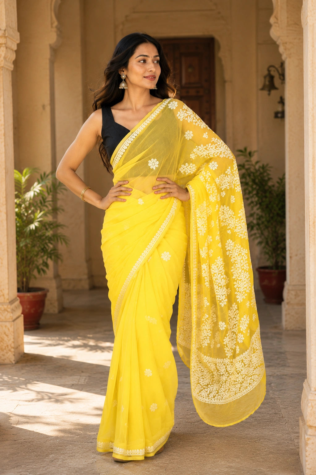 Lucknowi chikankari A woman wearing a bright yellow saree with a black sleeveless blouse, standing in a sunlit courtyard with potted plants and architectural columns. 