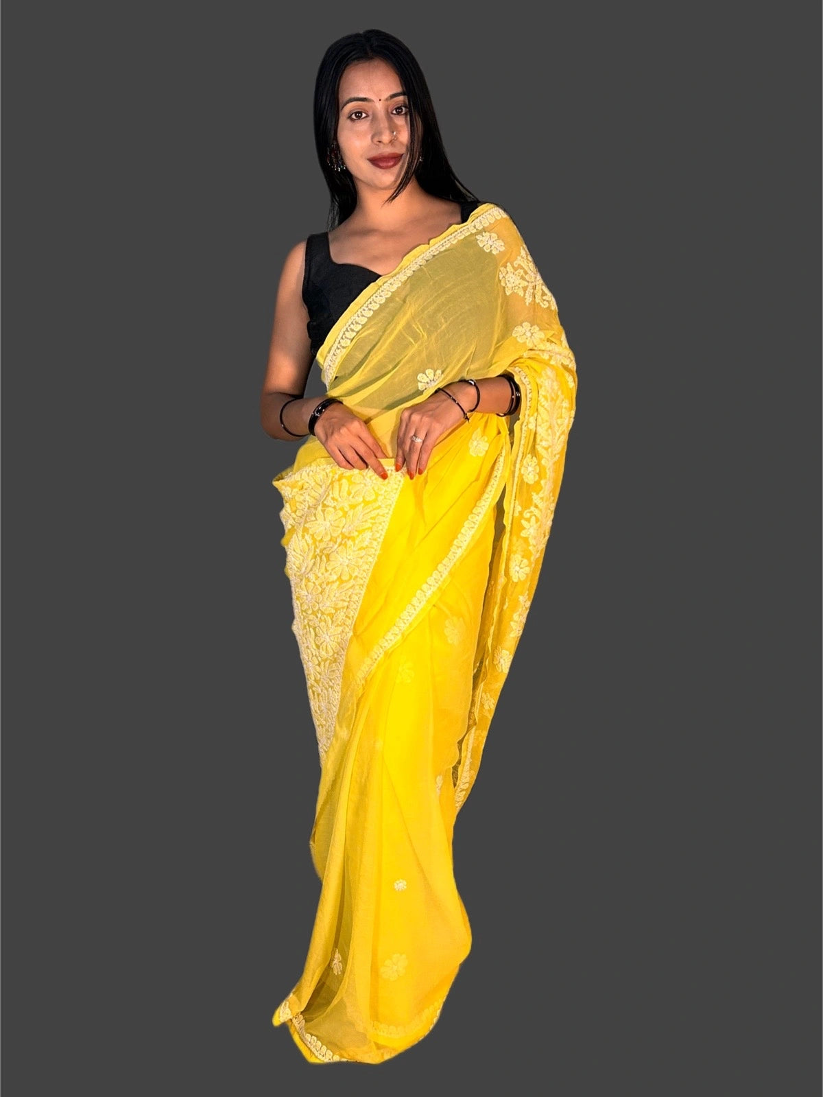 A woman modeling a bright yellow saree with white floral embroidery along the border, paired with a black sleeveless blouse, set against a solid dark gray background.