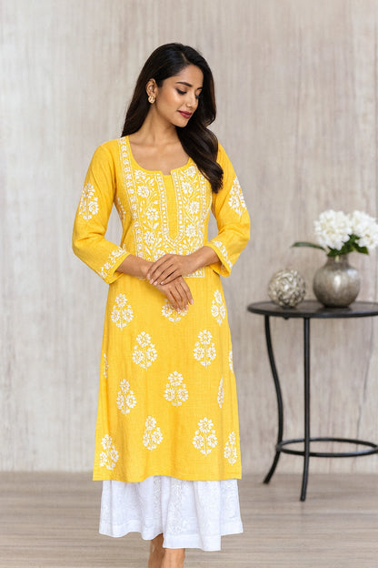 A woman modeling a bright yellow kurta with white floral embroidery and a white underskirt, standing in a styled indoor setting with a side table and flowers in the background.