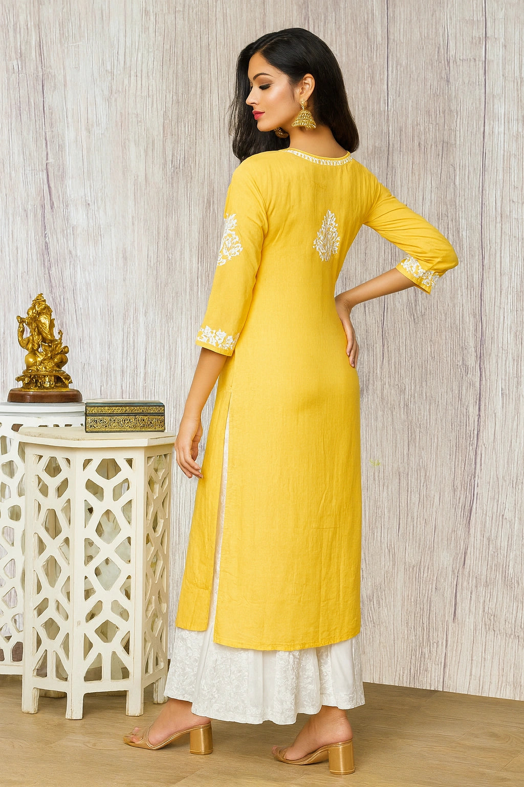 A woman wearing a light yellow embroidered kurta with white pants and beige block-heel sandals, shown from the back in a minimalist indoor setting.