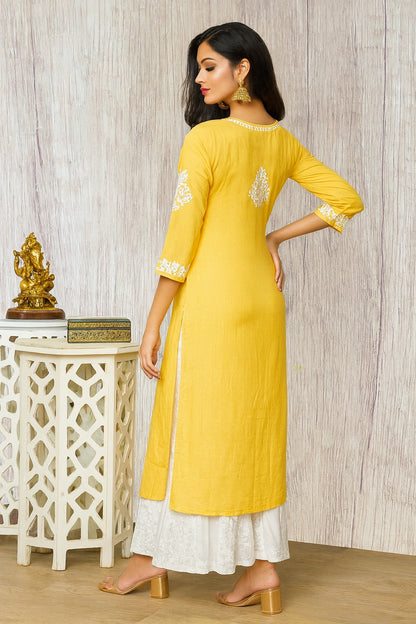 A woman wearing a light yellow embroidered kurta with white pants and beige block-heel sandals, shown from the back in a minimalist indoor setting.