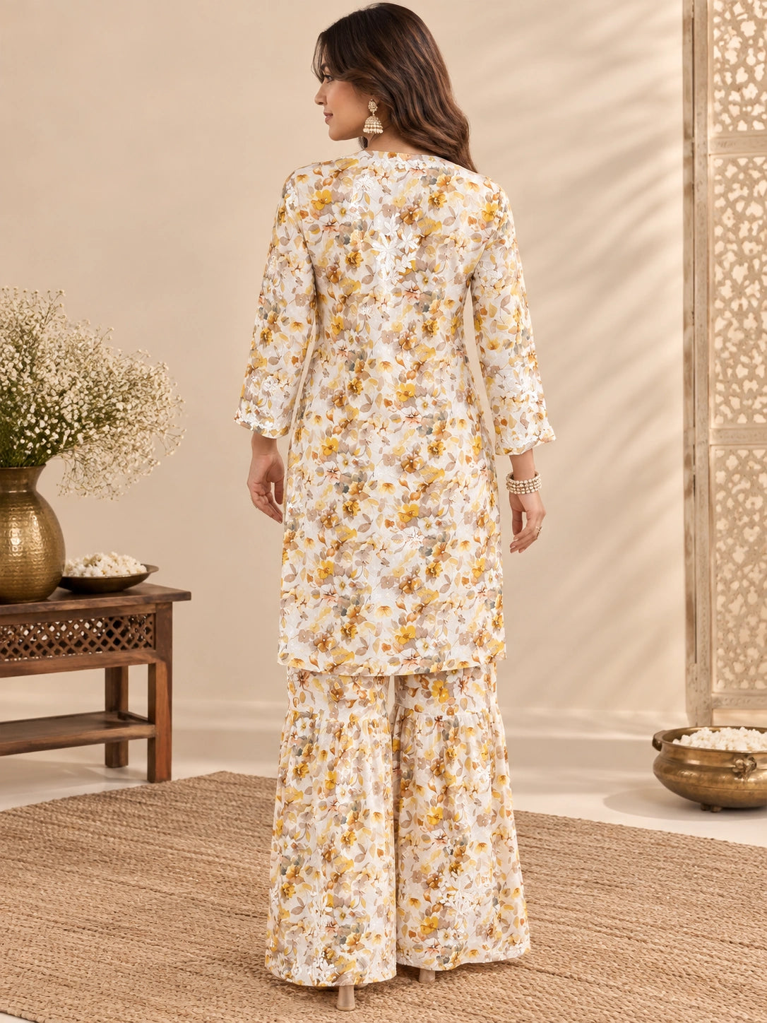  Lucknowi chikankari A woman stands with her back to the camera wearing a cream-colored two-piece kurta set with a yellow and orange floral pattern. The set includes a long-sleeved tunic and matching wide-leg pants, styled in a warm, neutral indoor setting with decorative plants and a wooden screen.