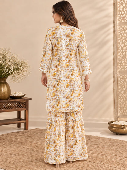  Lucknowi chikankari A woman stands with her back to the camera wearing a cream-colored two-piece kurta set with a yellow and orange floral pattern. The set includes a long-sleeved tunic and matching wide-leg pants, styled in a warm, neutral indoor setting with decorative plants and a wooden screen.