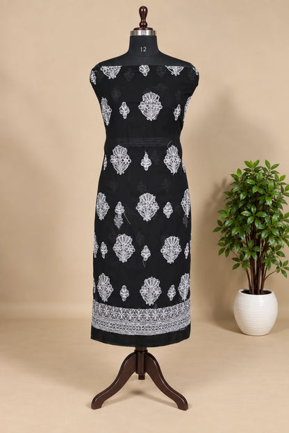 Lucknowi Chikankari A sleeveless black dress displayed on a wooden dress form mannequin, featuring white printed floral and geometric motifs with a decorative border at the hem, set against a beige background with a potted plant on the right.