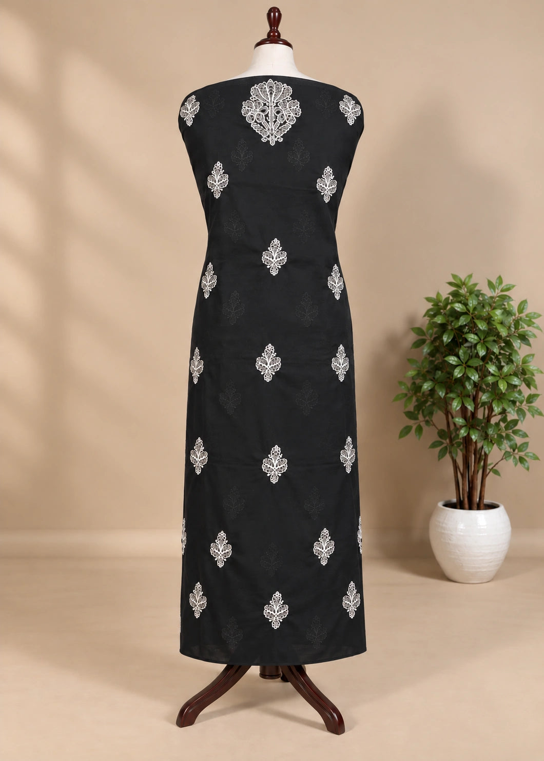 A sleeveless black dress displayed on a mannequin stand, featuring silver embroidered motifs down the front, set against a beige background with a potted plant on the right.
