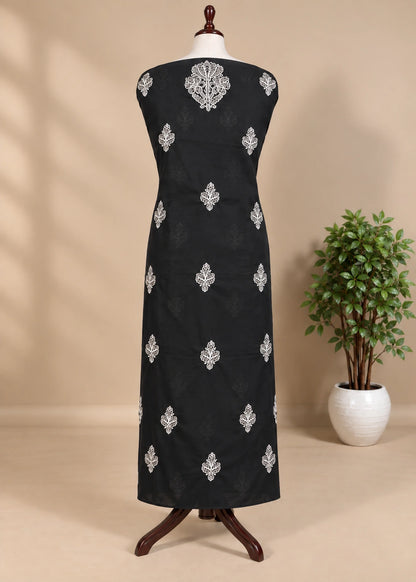A sleeveless black dress displayed on a mannequin stand, featuring silver embroidered motifs down the front, set against a beige background with a potted plant on the right.
