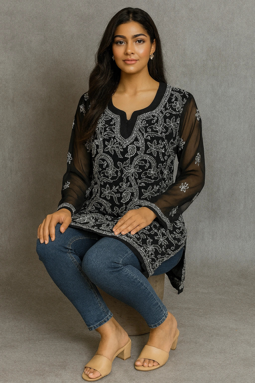 Woman wearing a black embroidered top and blue jeans sitting on a gray background