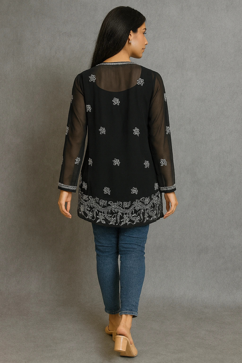 Woman wearing a black embroidered top with sheer sleeves and hem, standing against a gray background.