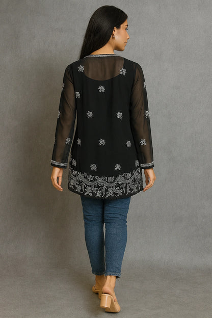 Woman wearing a black embroidered top with sheer sleeves and hem, standing against a gray background.