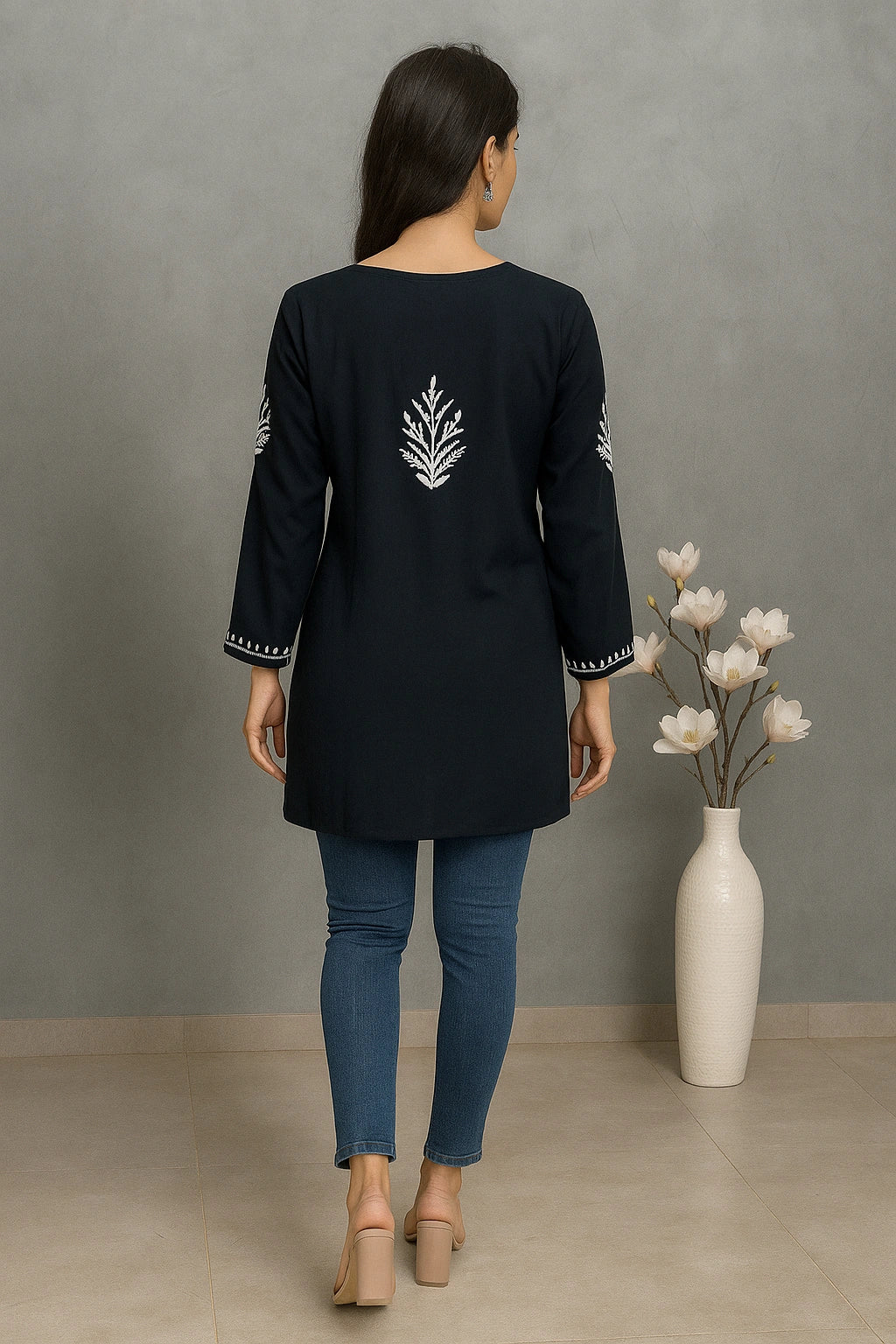 Woman wearing a black embroidered top and blue jeans standing against a gray wall with a white vase and flowers.