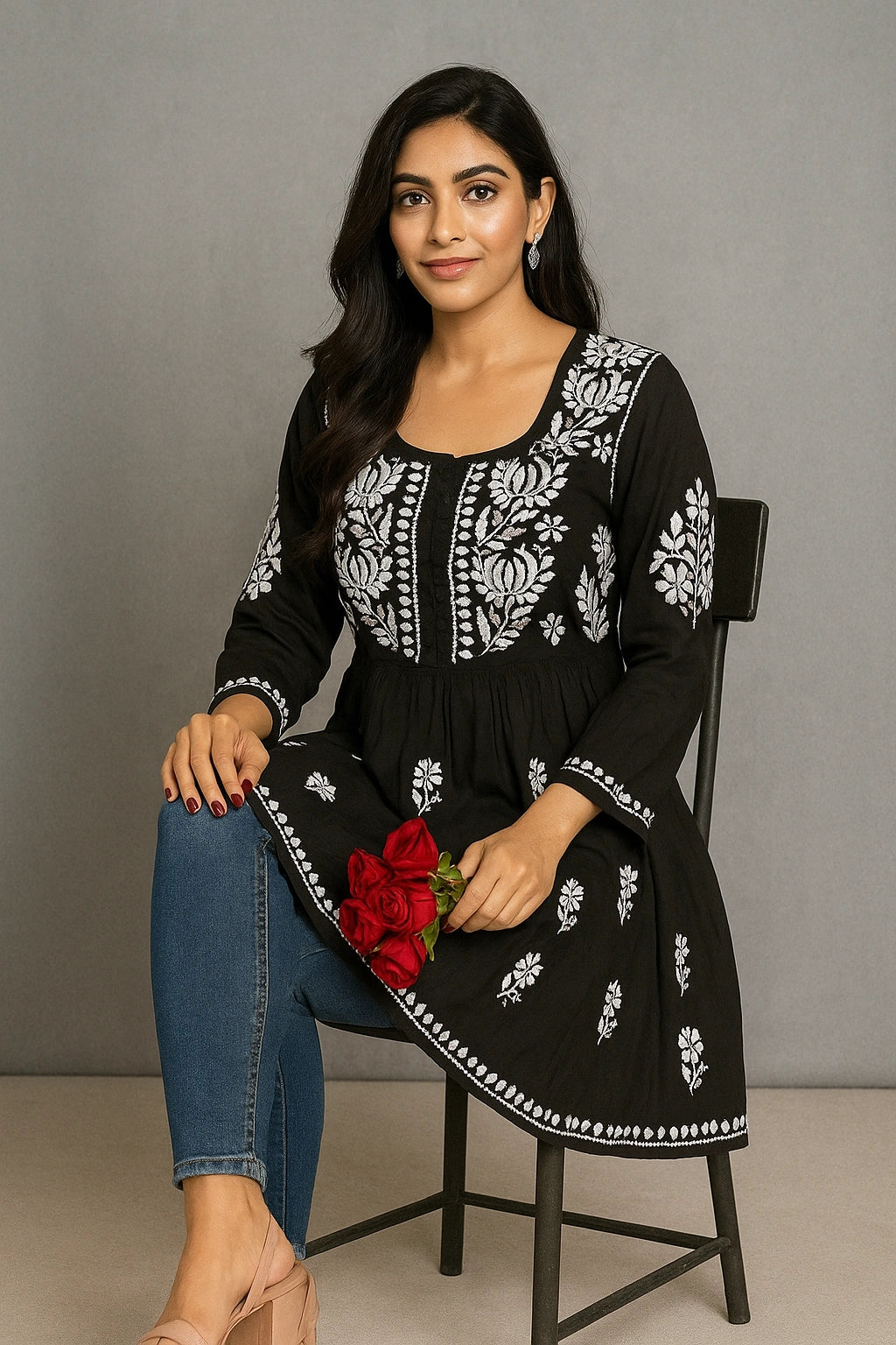 Woman wearing a black embroidered dress with white patterns, sitting on a chair against a gray background.