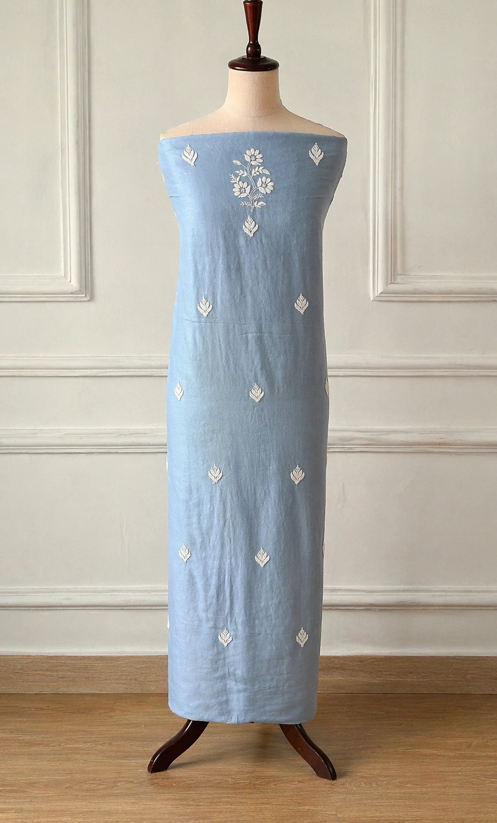 Light blue dress with white floral embroidery on a mannequin against a white paneled wall.