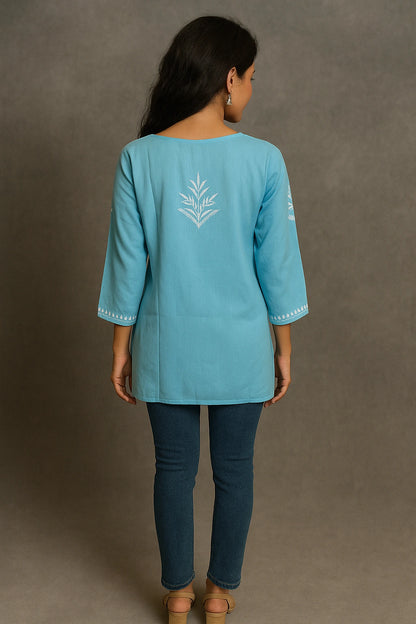 Woman wearing a light blue embroidered top and dark blue pants against a gray background