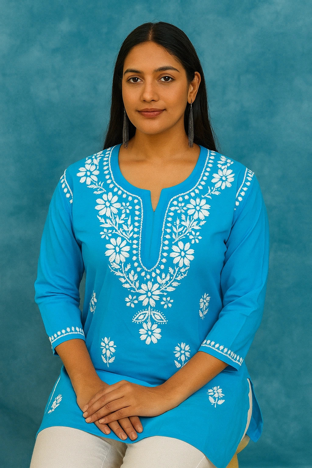 Woman sitting on a chair wearing a blue embroidered top and blue jeans against a gray background