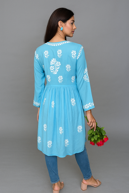Woman wearing a blue embroidered kurta with blue pants, holding green leaves and red flowers against a gray background.