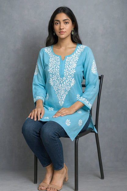 Woman sitting on a chair wearing a blue embroidered top and blue jeans against a gray background