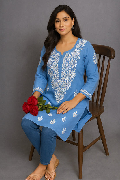Woman sitting on a chair wearing a blue embroidered top and blue jeans against a gray background