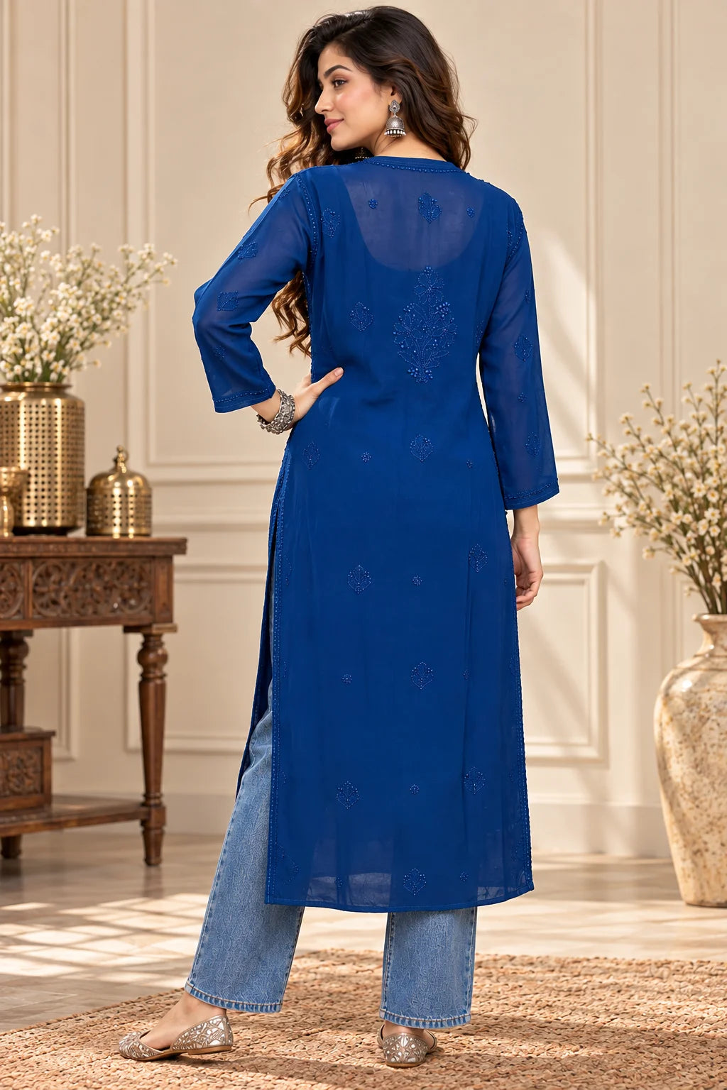 lucknowi chikankari A woman wearing a bright blue long kurta with light blue jeans and beige heels, photographed from the back in an elegant indoor setting with decorative vases and a wooden table.