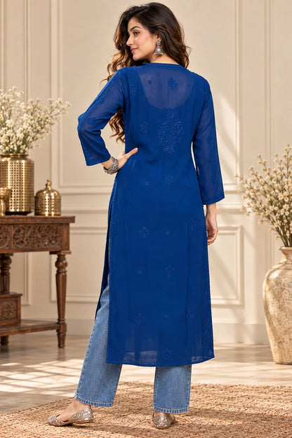 lucknowi chikankari A woman wearing a bright blue long kurta with light blue jeans and beige heels, photographed from the back in an elegant indoor setting with decorative vases and a wooden table.