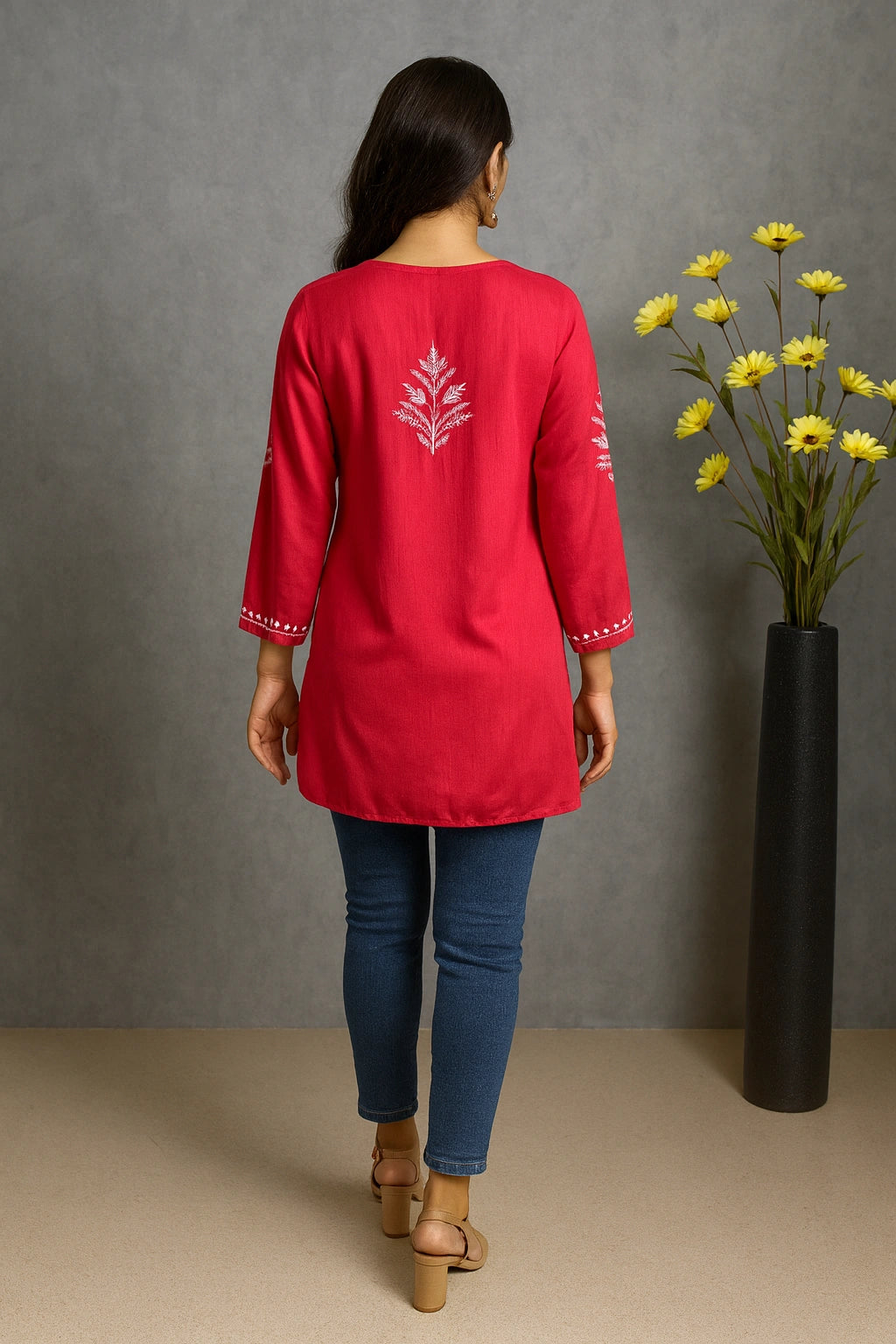 Woman wearing a red top with floral embroidery, standing against a gray wall with a vase of yellow flowers.