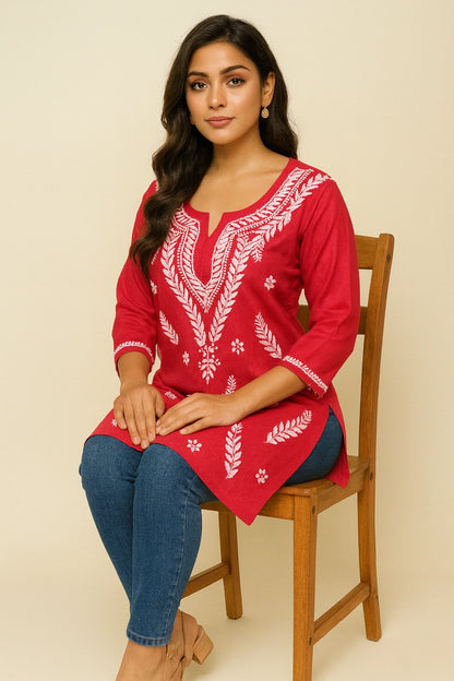 Woman wearing a red embroidered top sitting on a wooden chair against a beige background
