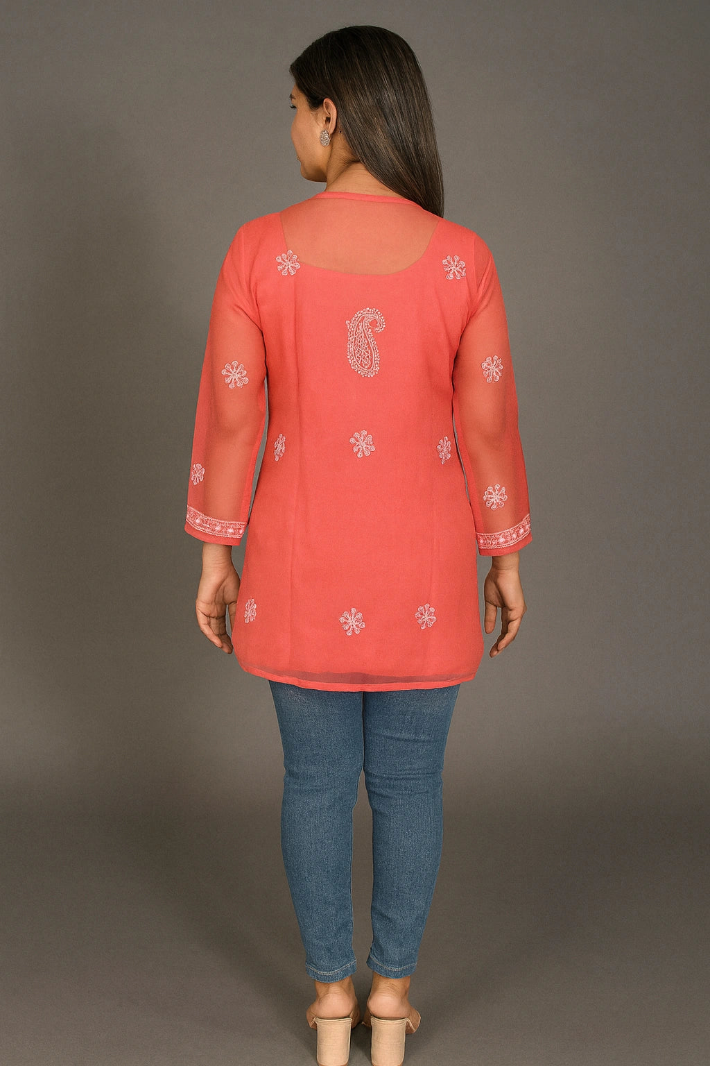 Woman wearing a coral pink embroidered top and blue jeans against a gray background