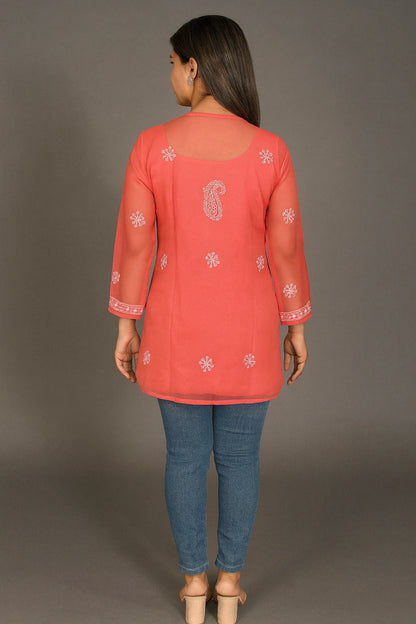 Woman wearing a coral pink embroidered top and blue jeans against a gray background
