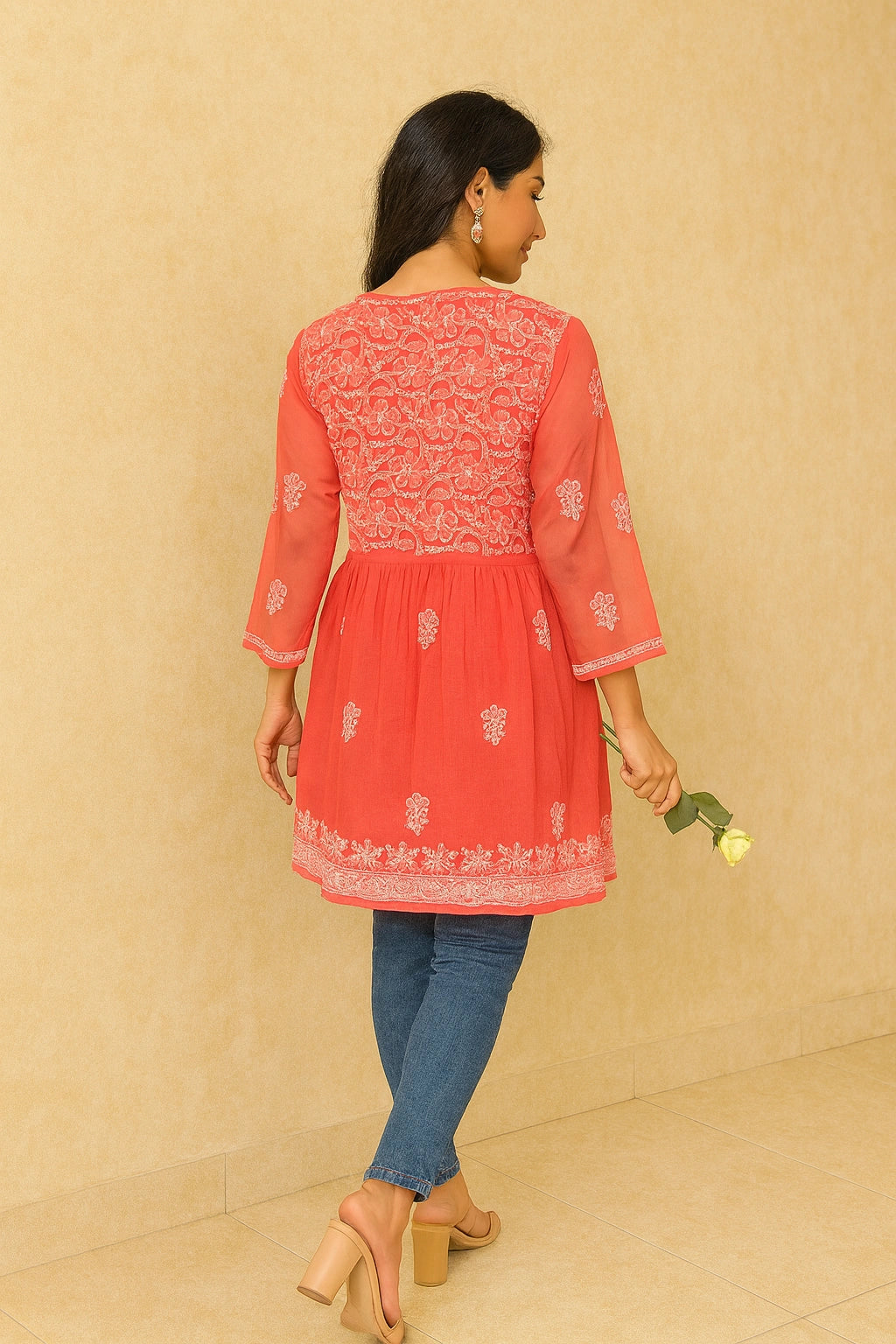 Woman wearing a coral embroidered top with blue jeans against a beige background