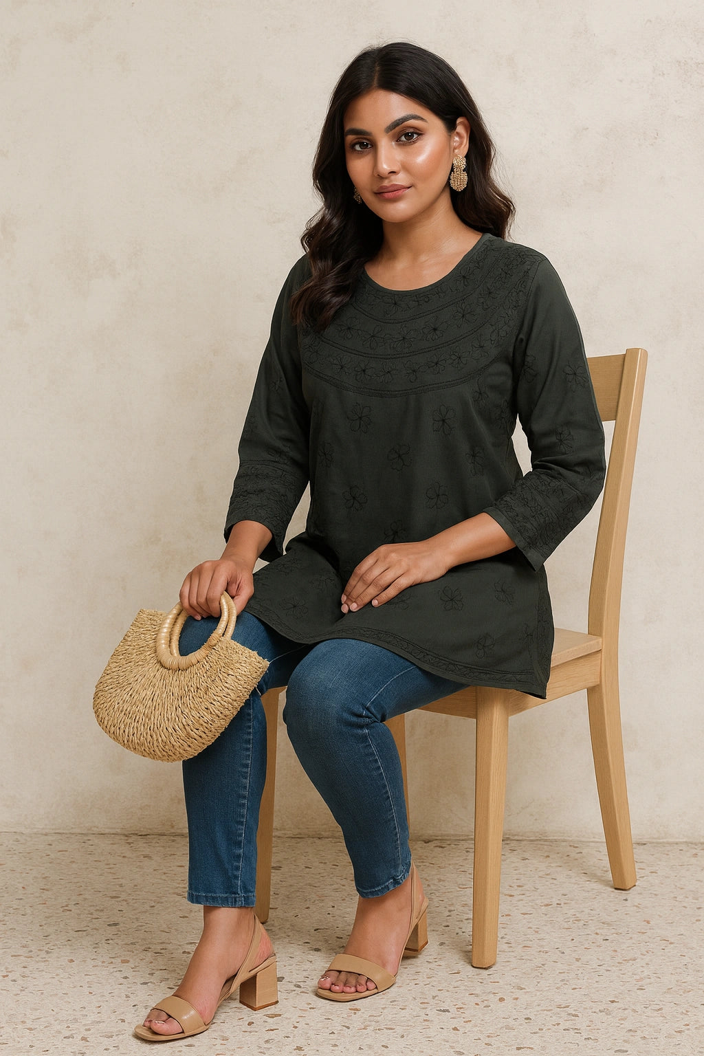 lucknowi chikankari Woman sitting on a chair holding a woven handbag, wearing a dark green top and blue jeans.