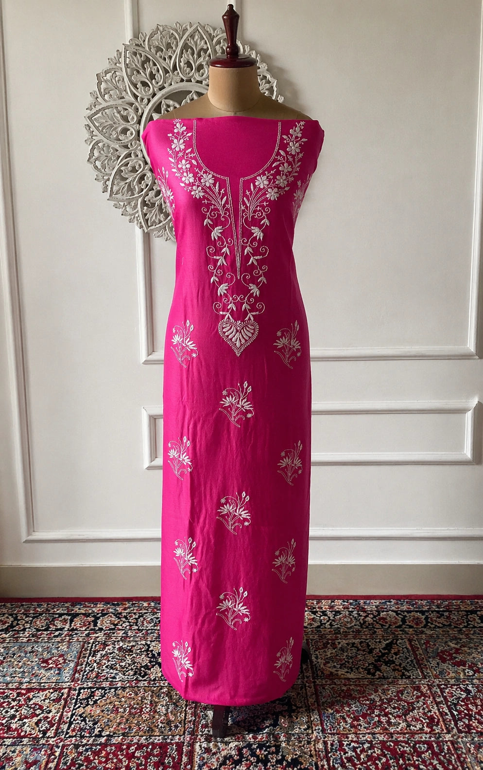 Pink dress with white floral embroidery on a mannequin against a decorative wall.