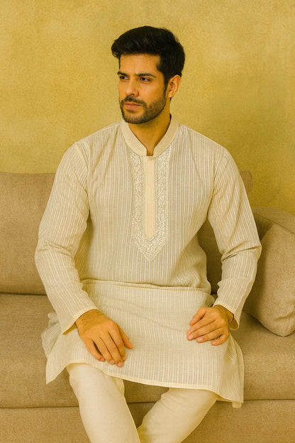 Man wearing a traditional lucknowi chikankari kurta sitting on a beige couch against a beige wall.