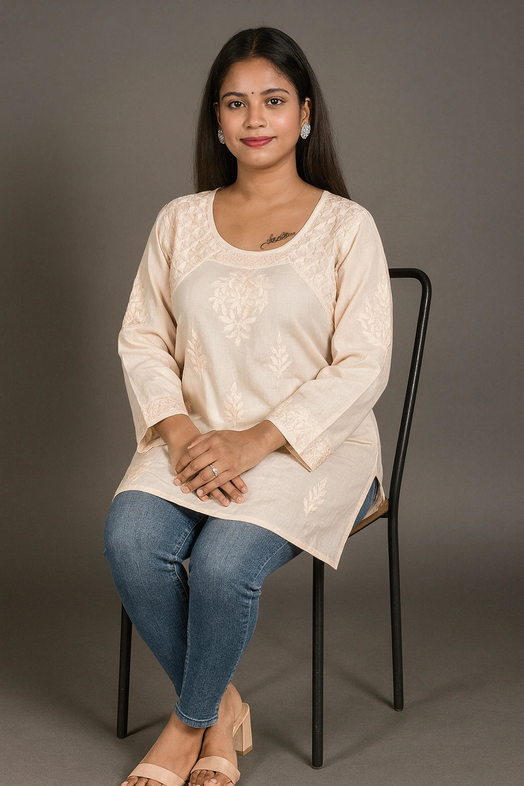 lucknowi chikankari Woman sitting on a chair wearing a beige top and blue jeans against a gray background