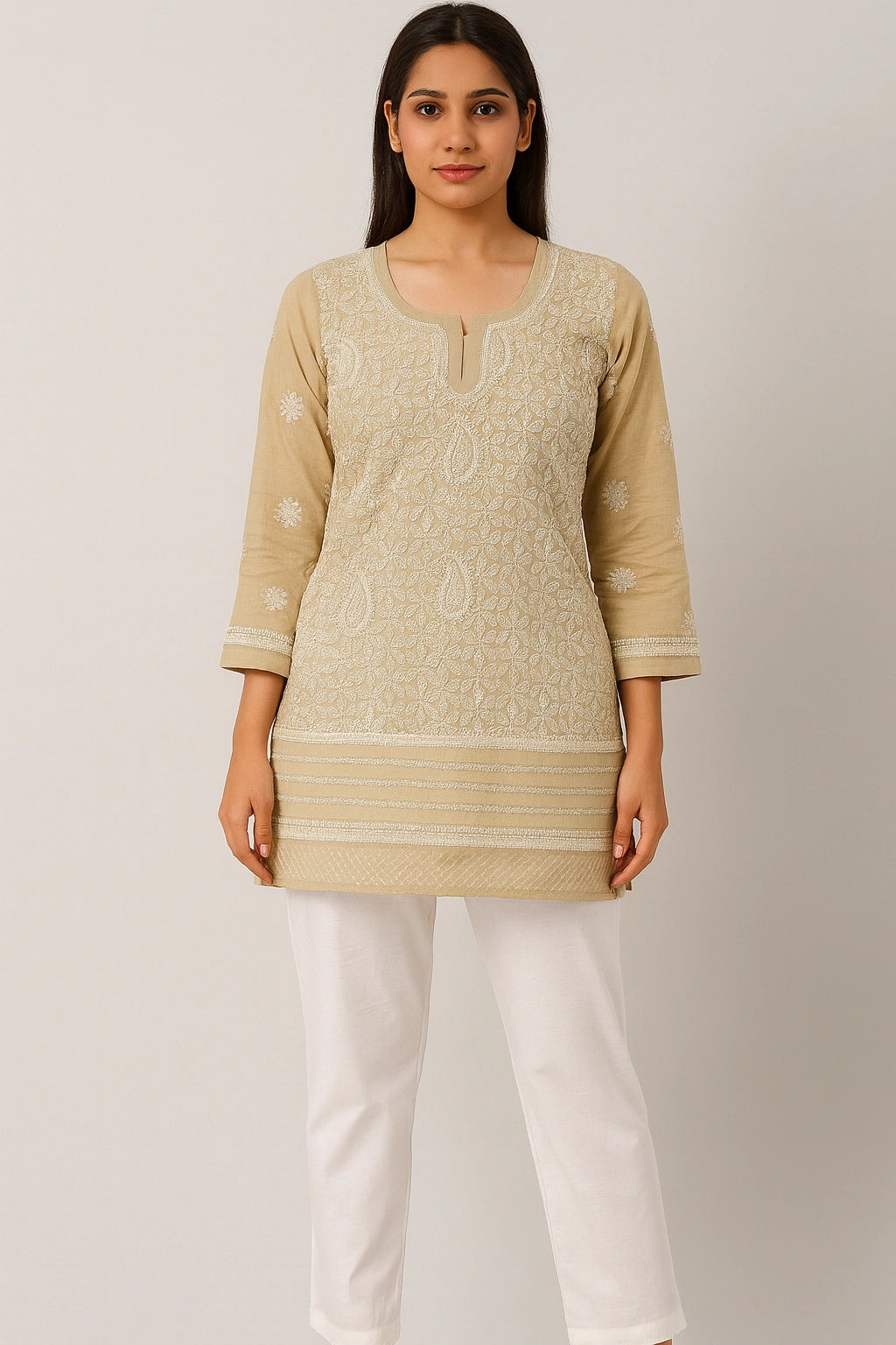 Woman wearing a beige embroidered top and white pants on a plain background