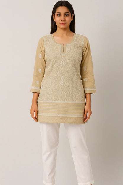 Woman wearing a beige embroidered top and white pants on a plain background