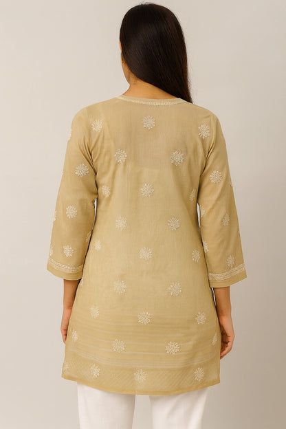 Woman wearing a beige embroidered top and white pants on a plain background