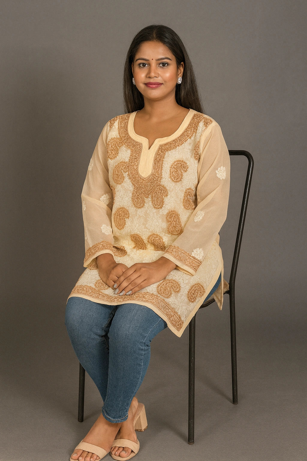 Woman sitting on a chair wearing a beige embroidered top and blue jeans against a gray background