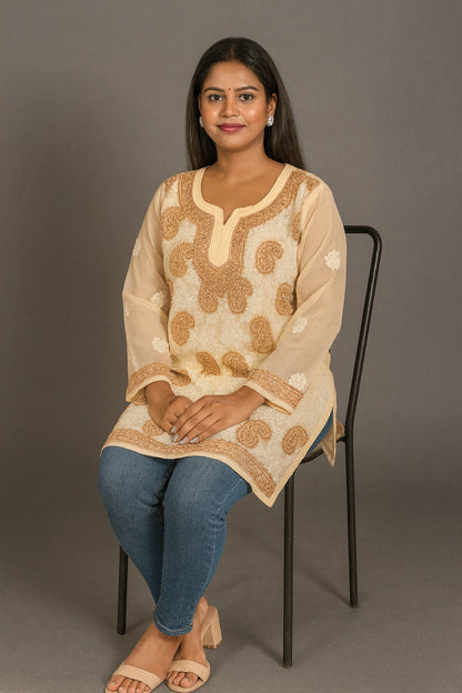 Woman sitting on a chair wearing a beige embroidered top and blue jeans against a gray background