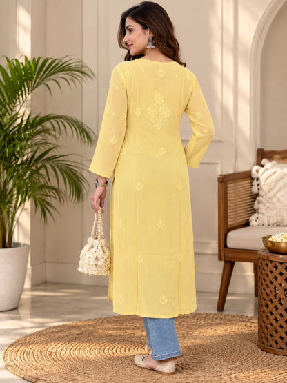 A woman stands outdoors wearing a long fawn-colored embroidered kurta with three-quarter sleeves, paired with light blue jeans and beige heels. The background features a warm-toned patio with potted plants and a wooden bench.